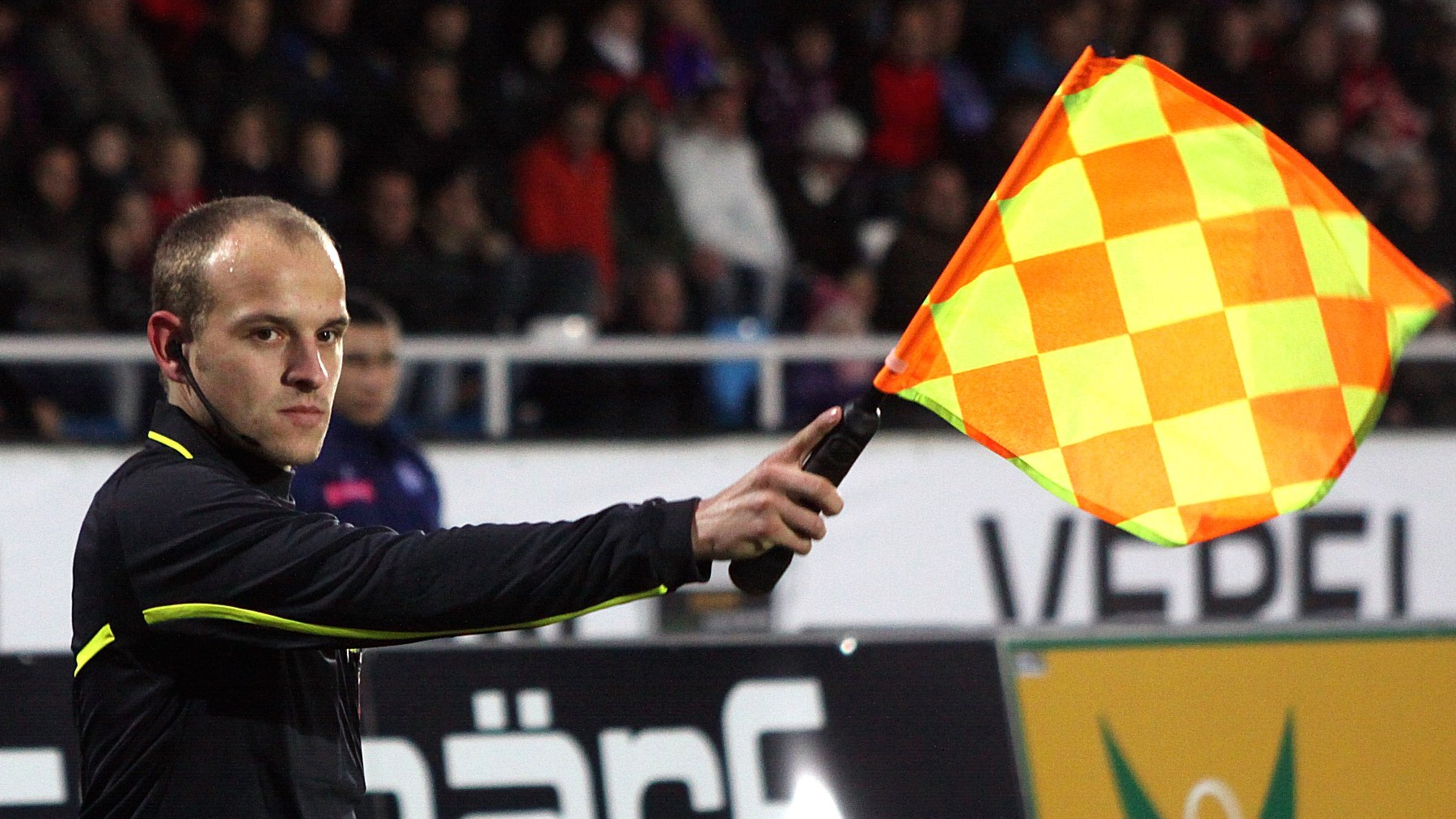Assisterande domare på sidlinjen med lyft gul-orange flagga som signalerar offside i en fotbollsmatch